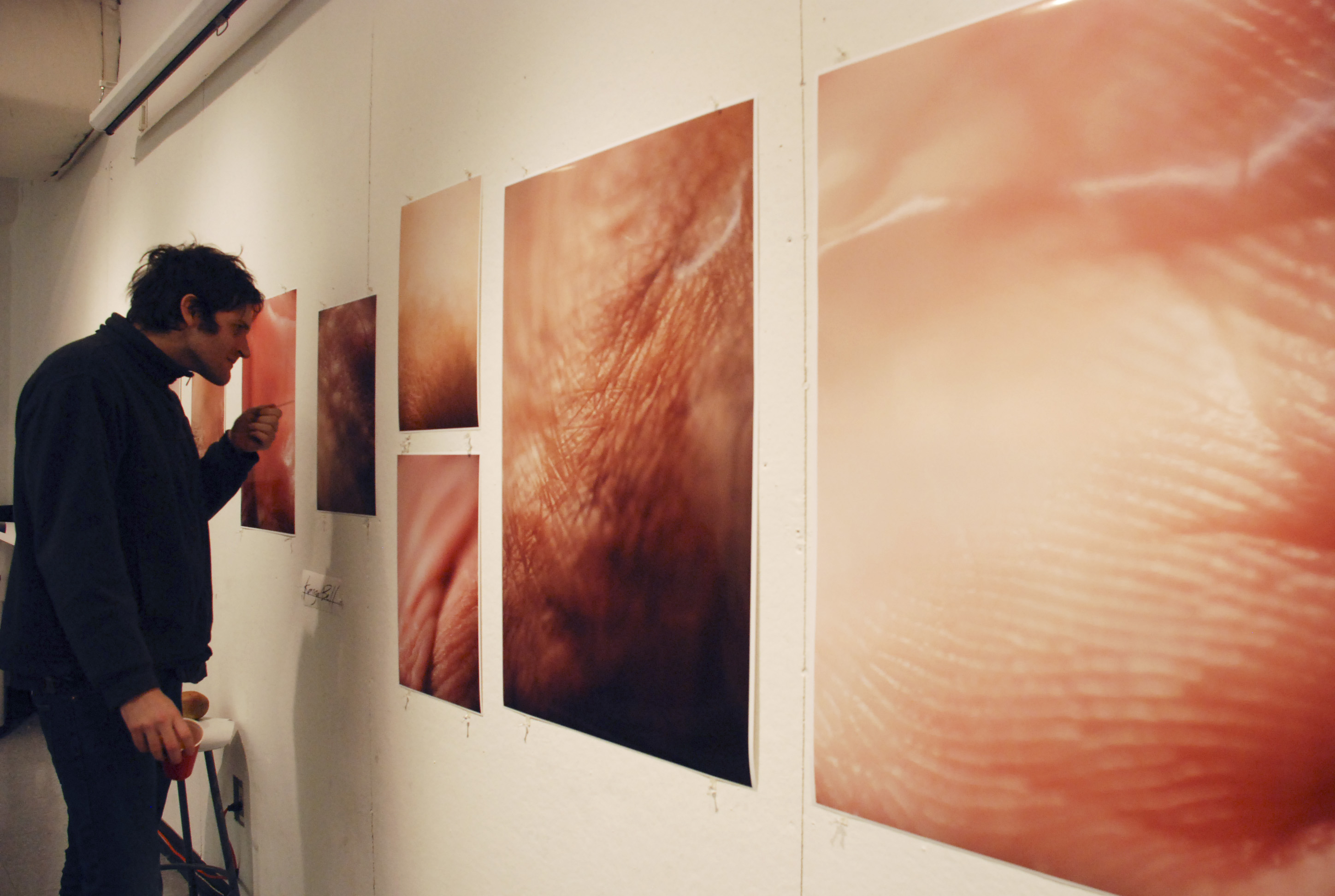 Large glossy closeup photos of human flesh on a white wall, with a person inspecting one of the photos and holding a plastic cup.